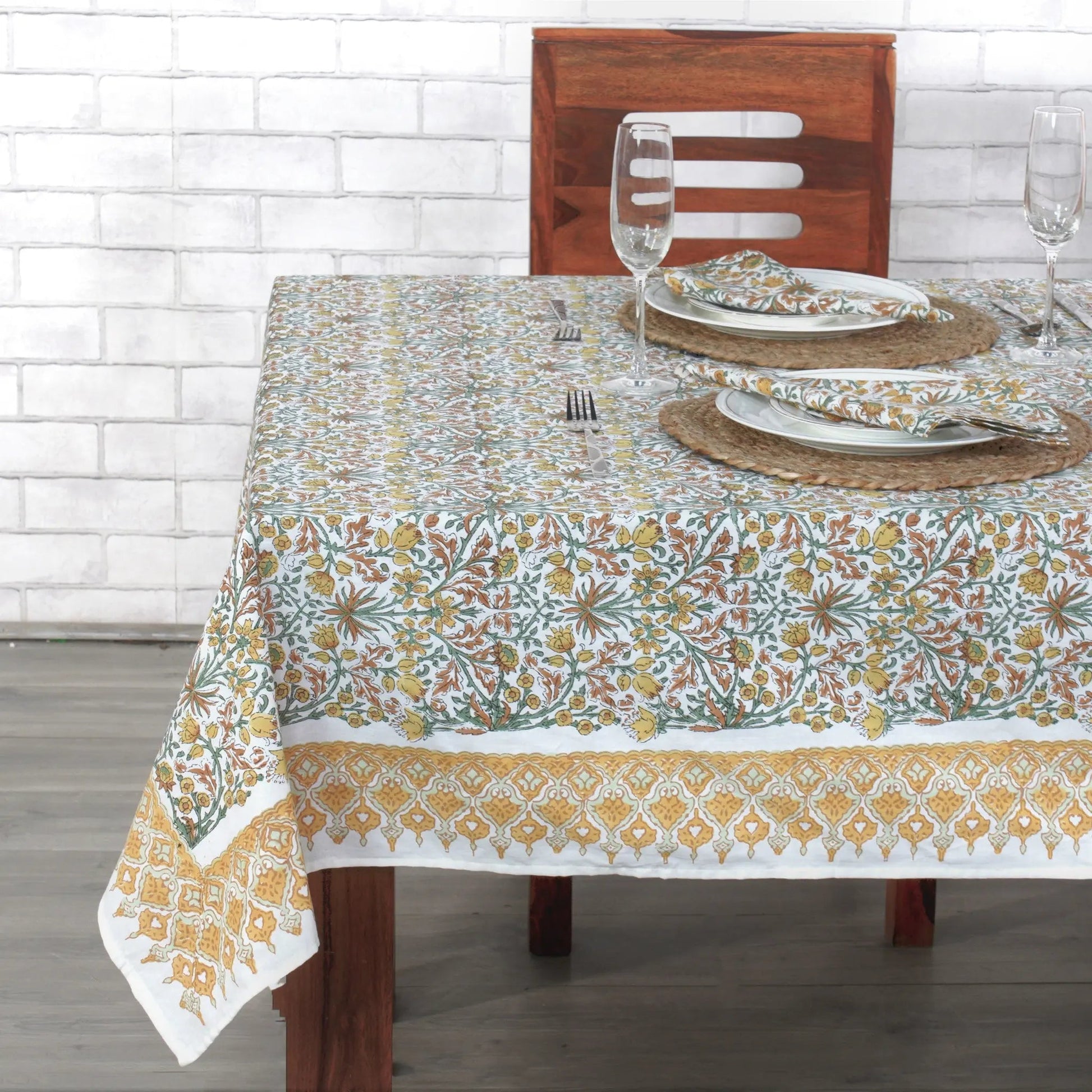 Dining table set with a floral patterned tablecloth against a white brick wall.