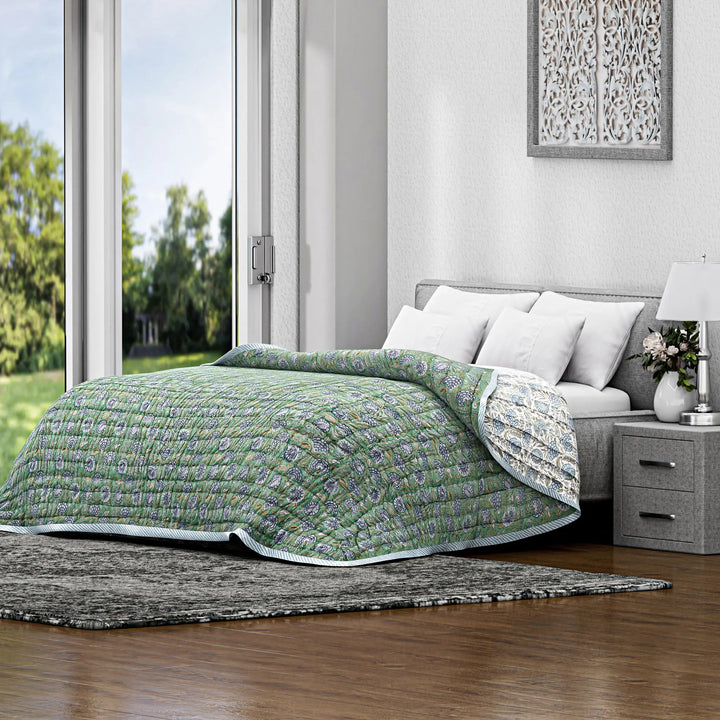 Bed with green quilt in a bright bedroom with large windows.