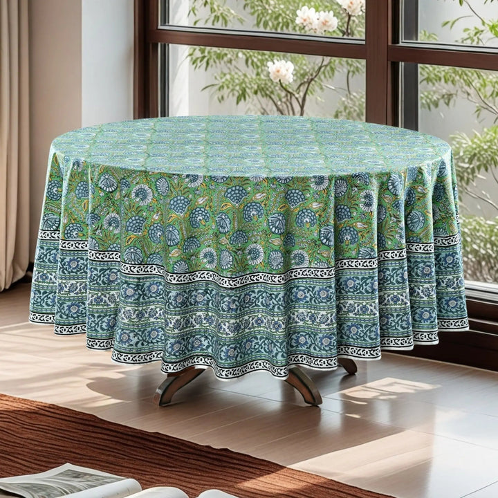 Round table with a green floral patterned tablecloth in a bright room with windows.
