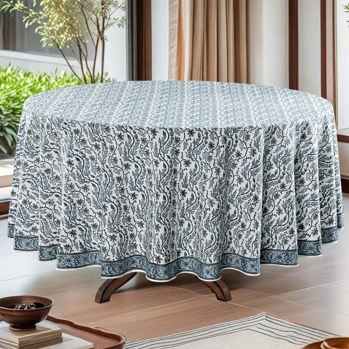 Round table with a patterned tablecloth in a room with plants and curtains.