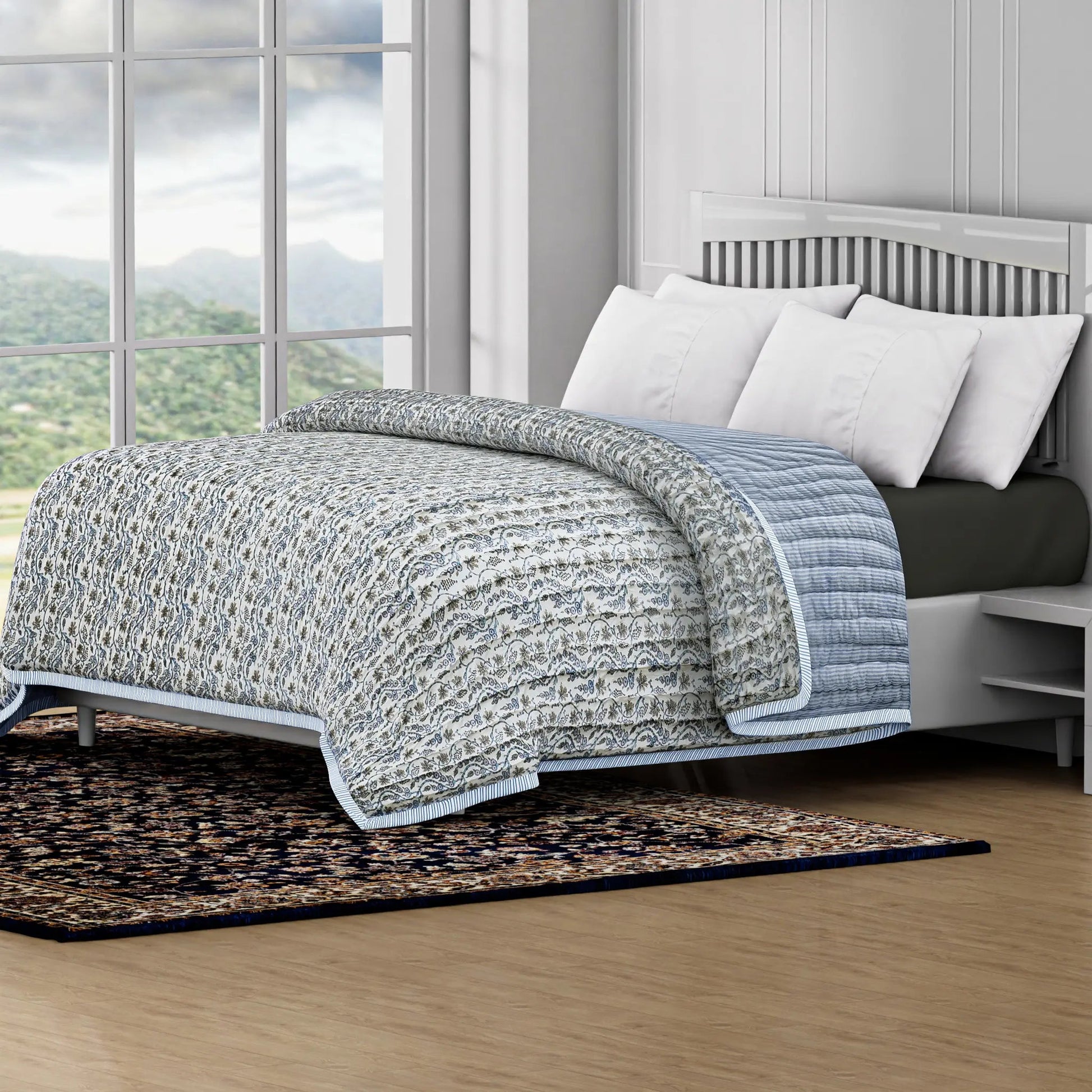 Bed with a textured quilt and white pillows in a room with large windows showing a scenic view.