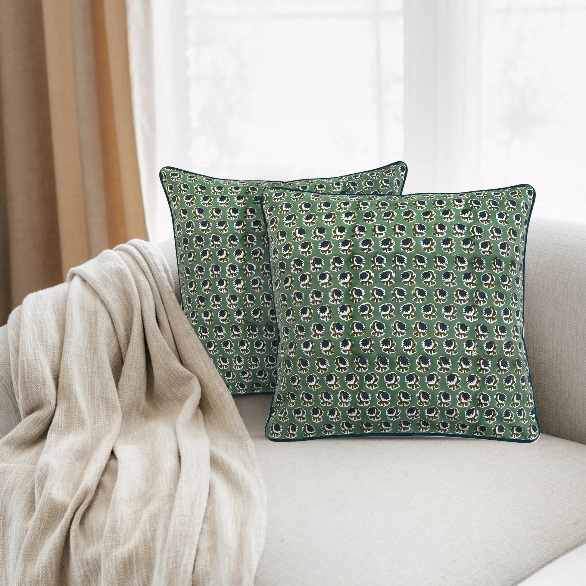 Two green patterned pillows on a beige sofa with a neutral curtain in the background