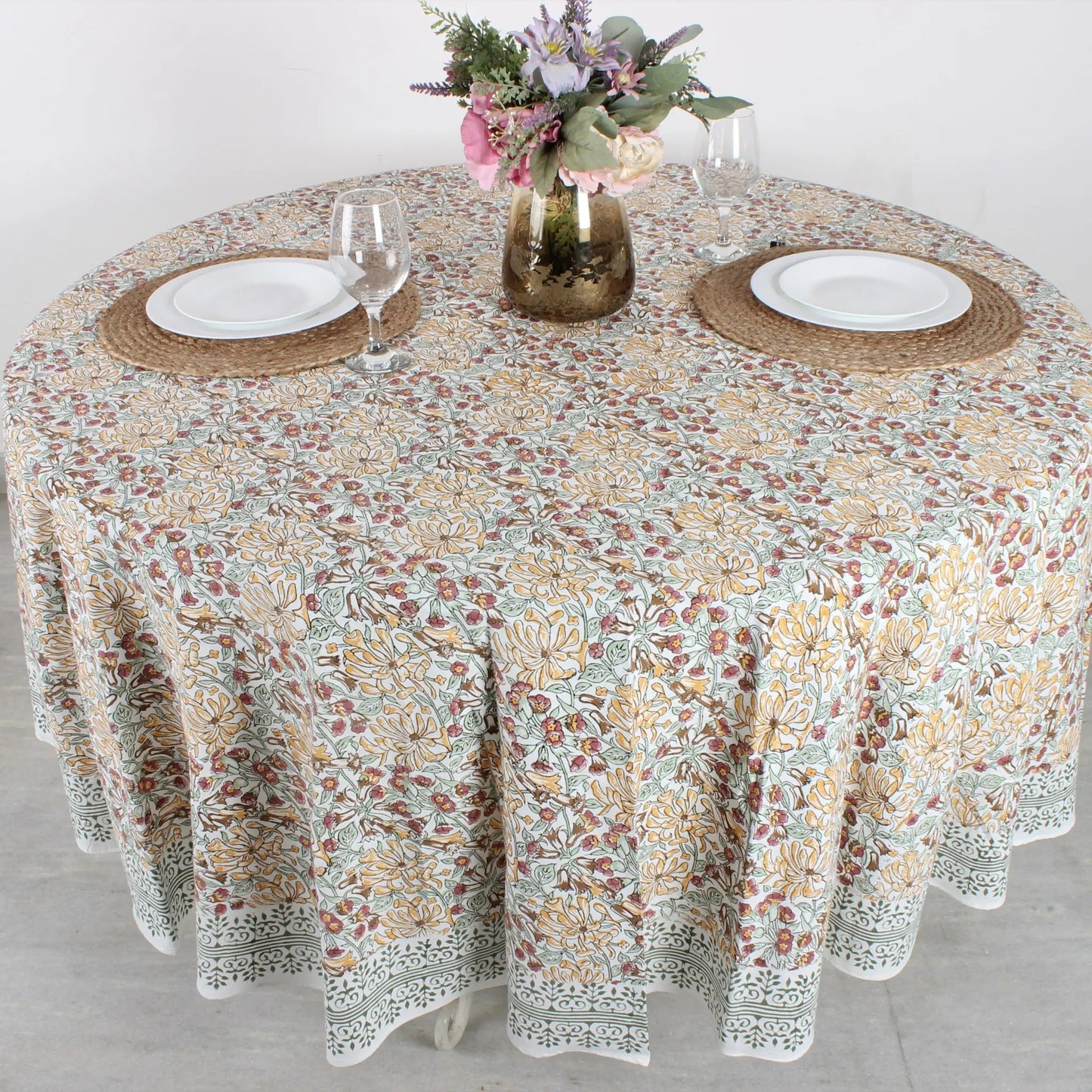 Round table with floral patterned tablecloth, plates, and glasses on a light background