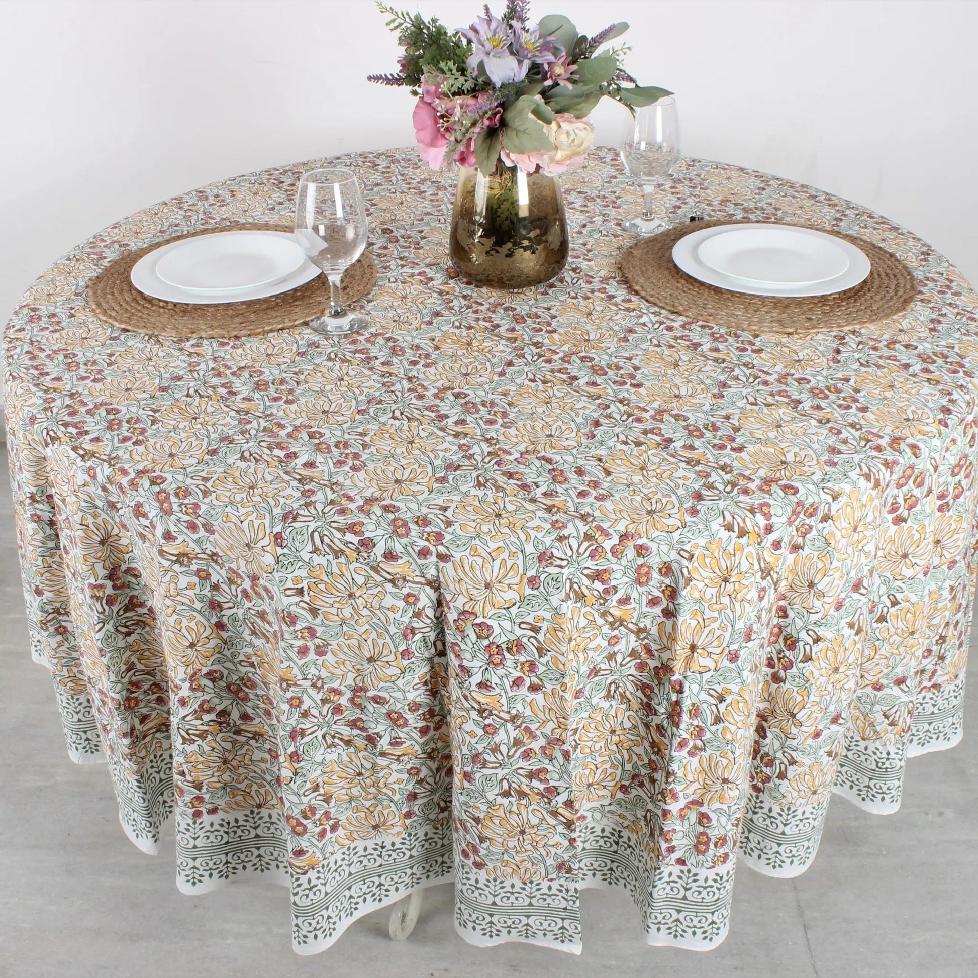 Round table with floral patterned tablecloth, plates, and glasses on a light background