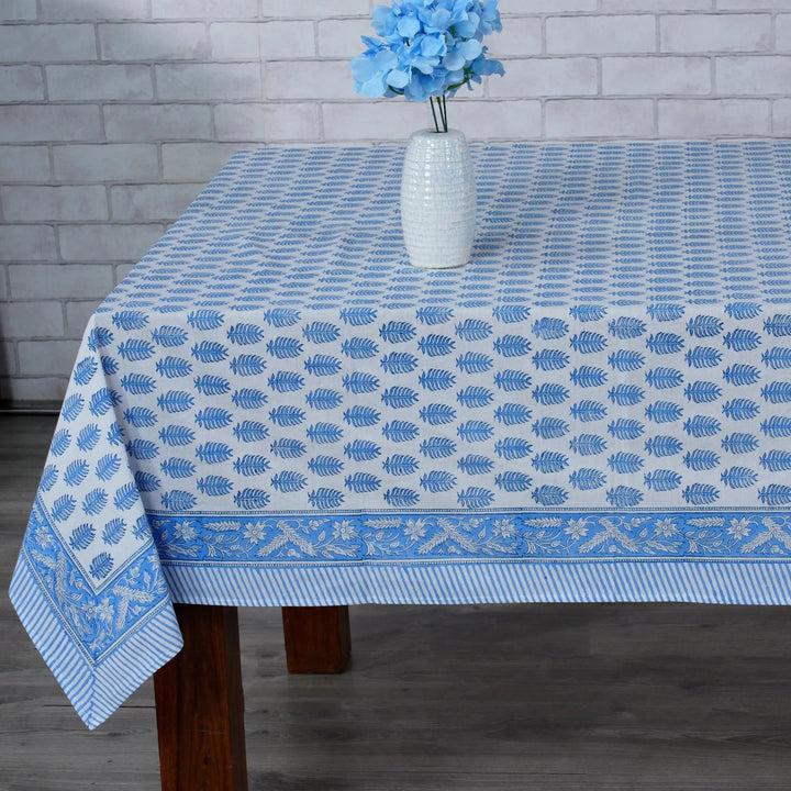 Blue patterned tablecloth on a wooden table with a vase of blue flowers against a gray brick wall.