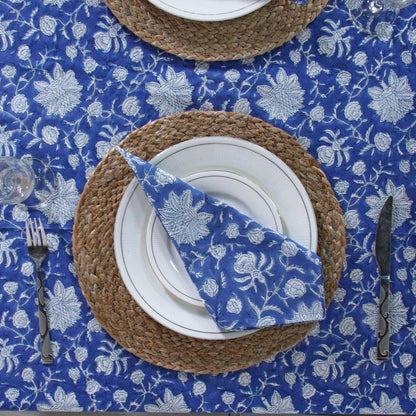 Table setting with blue floral tablecloth, woven placemat, white plates, and silverware.