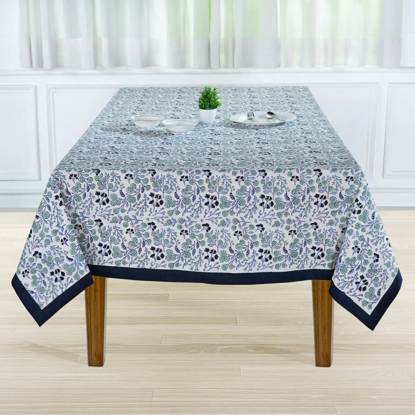 Tablecloth with floral pattern on a wooden table in a room with white walls and curtains.