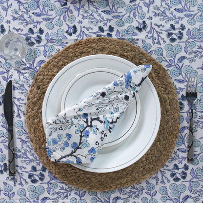 Table setting with a woven placemat, white plates, and a floral-patterned napkin on a blue and white tablecloth.