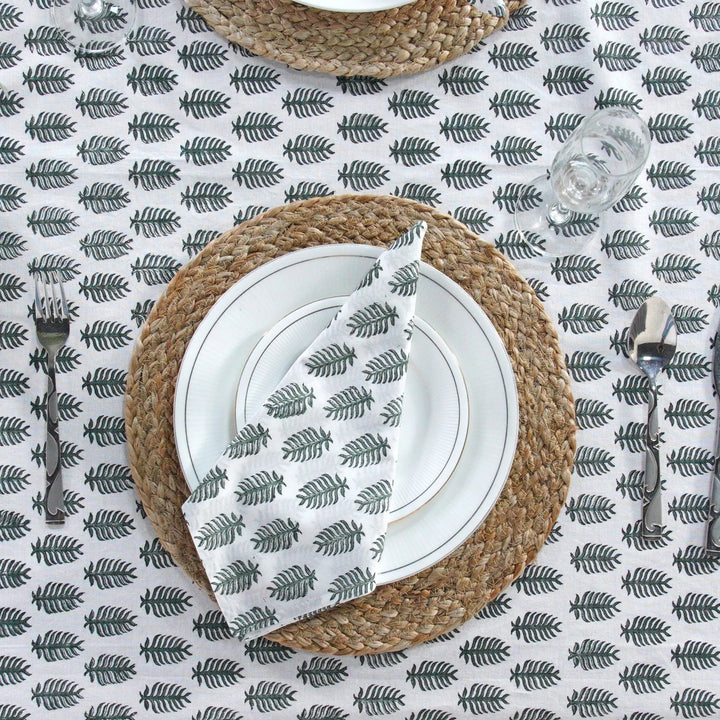 Table setting with woven placemat, white plates, and silverware on a leaf-patterned tablecloth.