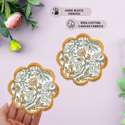 Two floral coasters with gold edges on a light pink background, held by a hand.