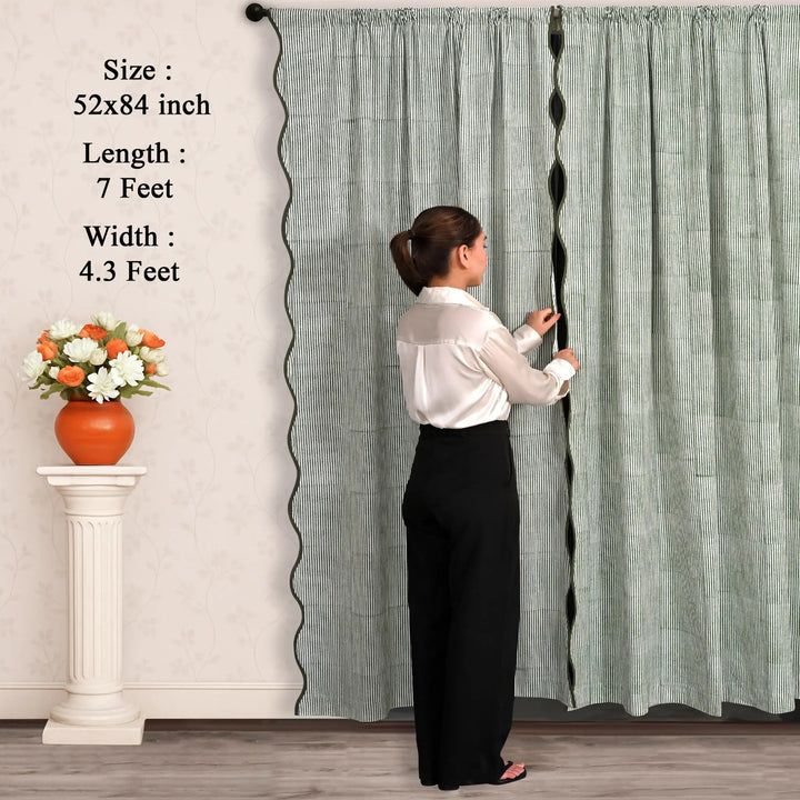 Woman adjusting a long curtain with text displaying dimensions, next to a small table with flowers.