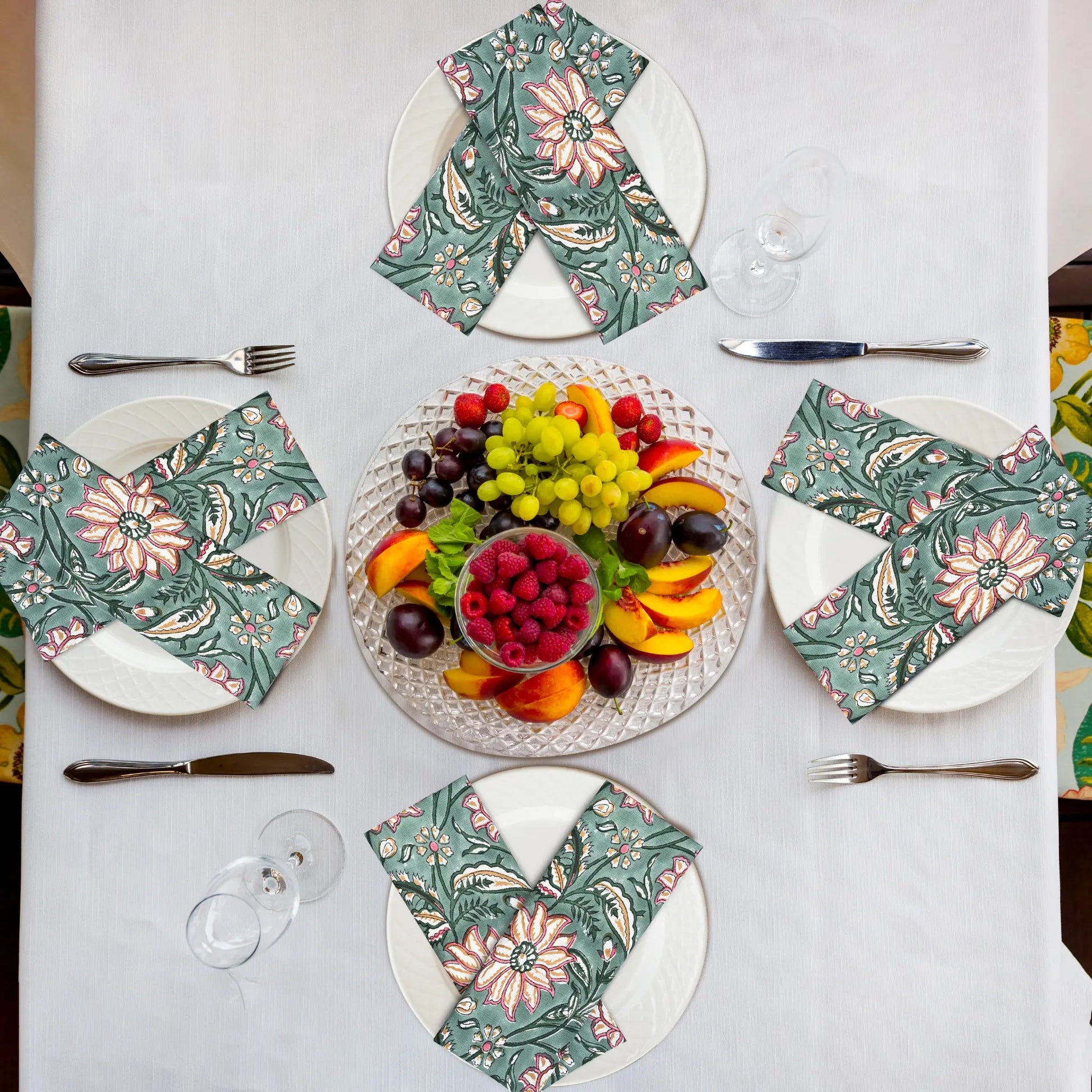 Table setting with floral napkins, fruit platter, and cutlery on a white tablecloth.