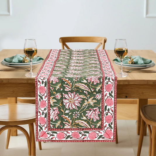 Decorative floral table runner on a wooden table with glasses and plates.