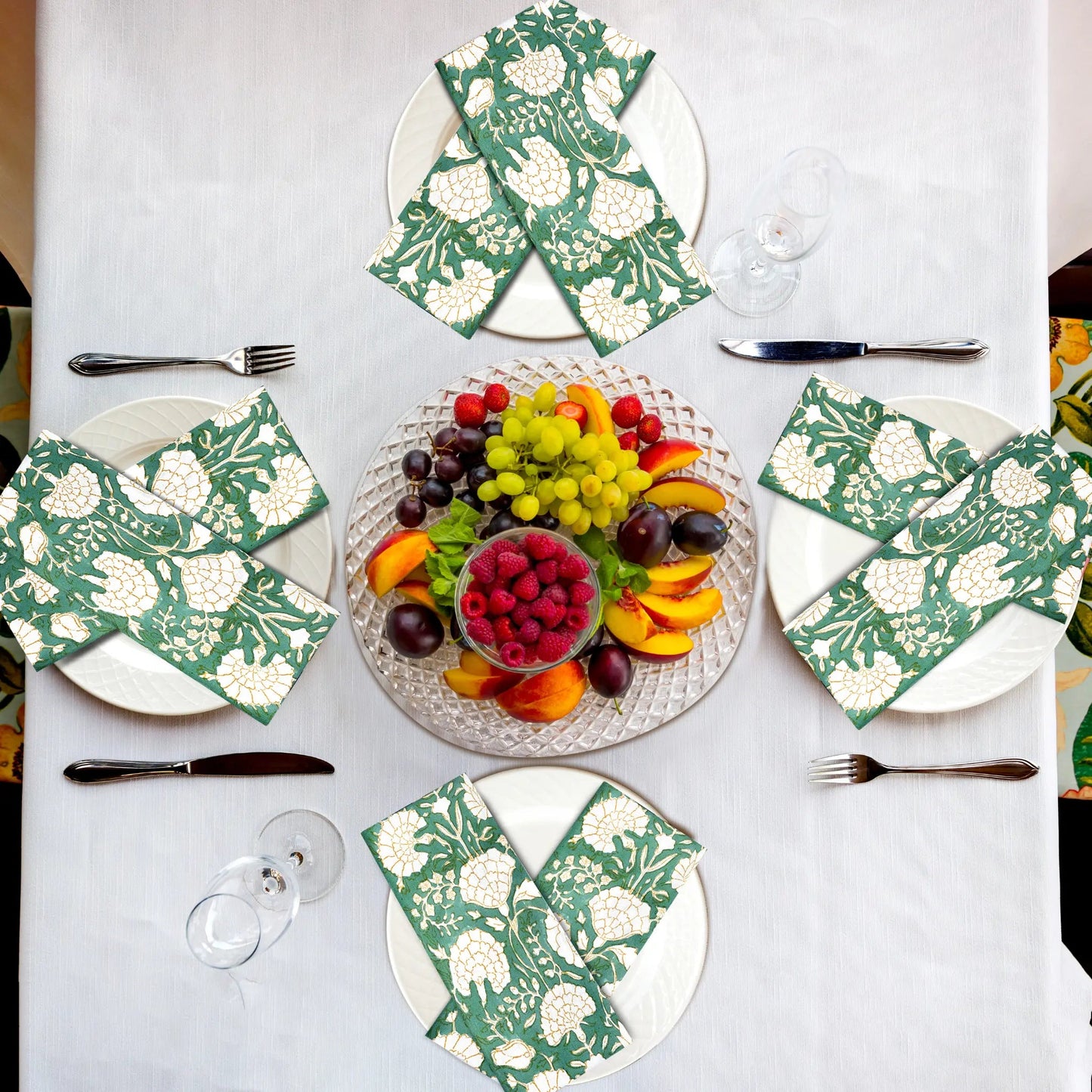 Table setting with floral napkins, fruit platter, and cutlery on a white tablecloth.