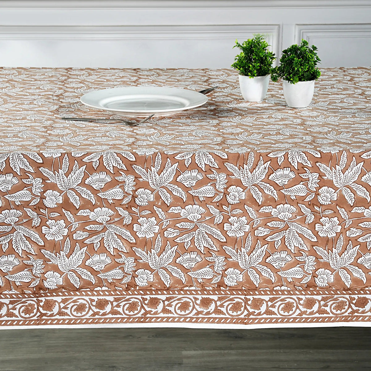 Table setting with a floral patterned tablecloth, white plate, and potted plants.