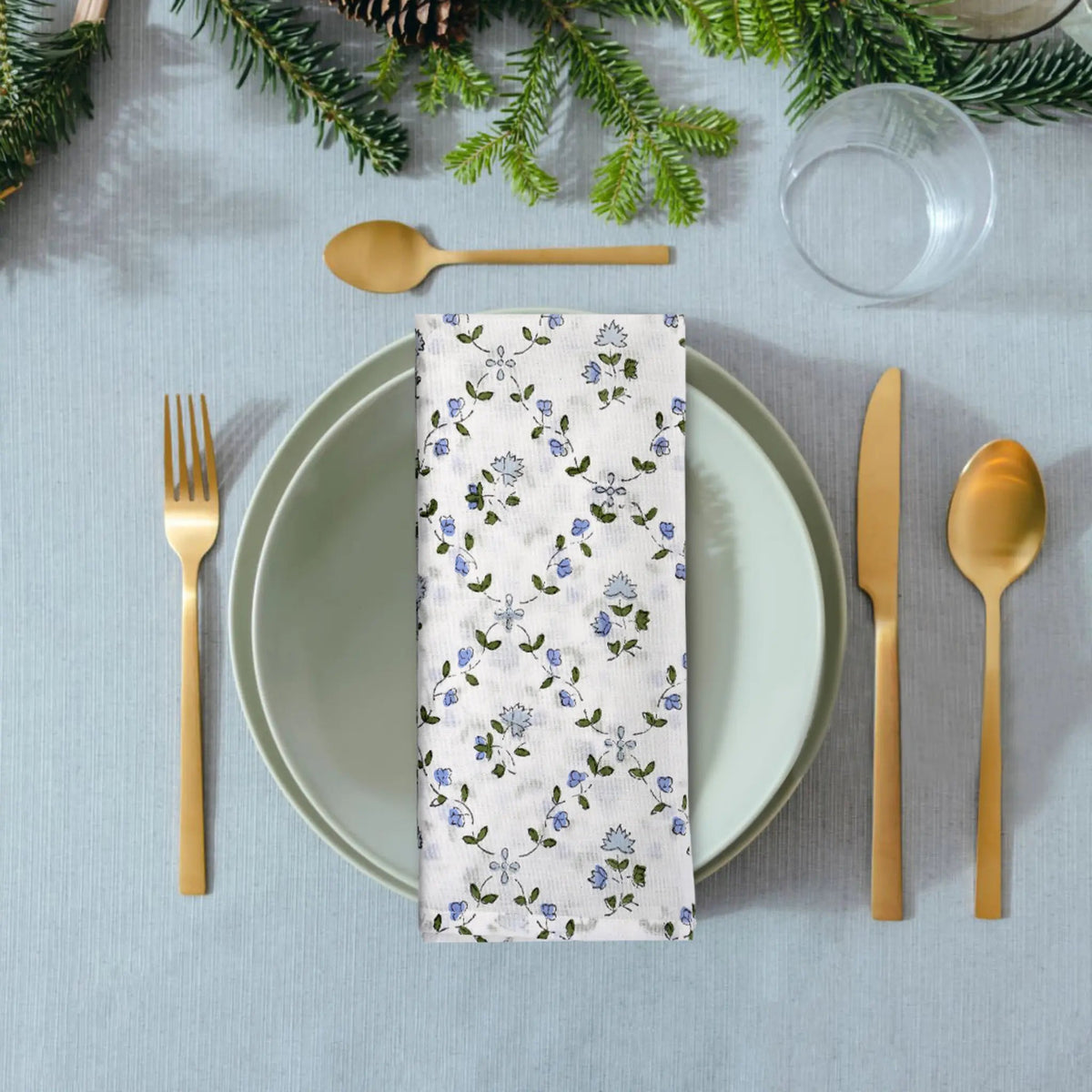 Table setting with floral napkin, gold cutlery, and greenery on a light blue tablecloth.