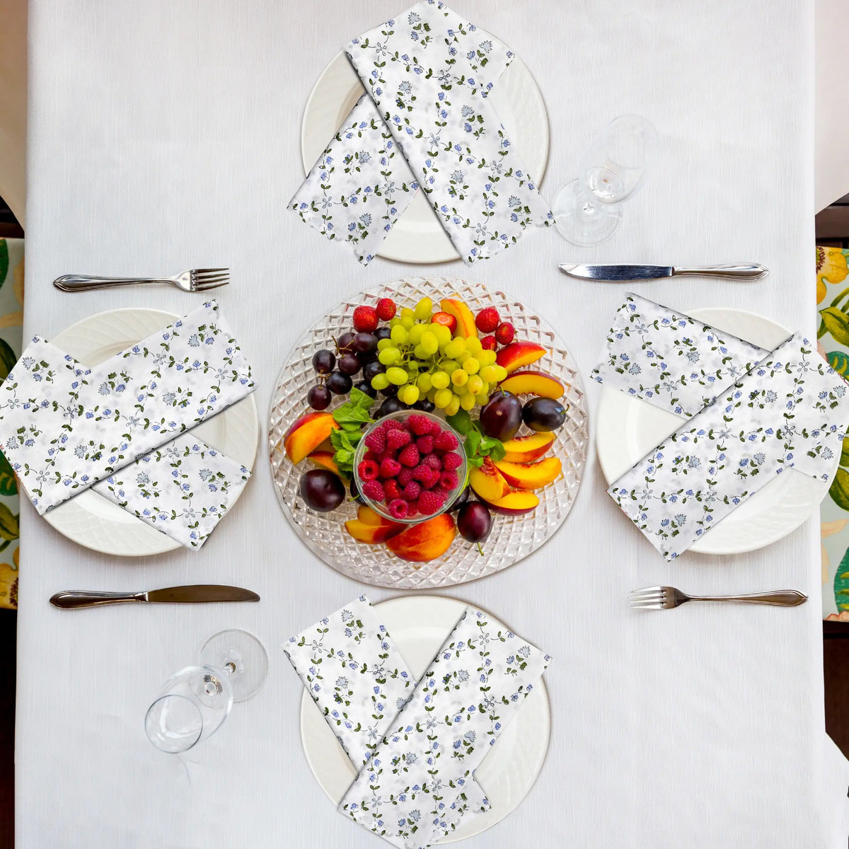 Table setting with floral plates, fruit platter, and cutlery on a white tablecloth.