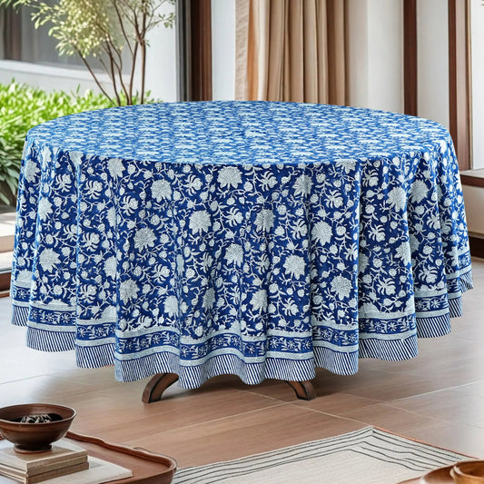 Round table covered with a blue floral patterned tablecloth in a room with plants and curtains.
