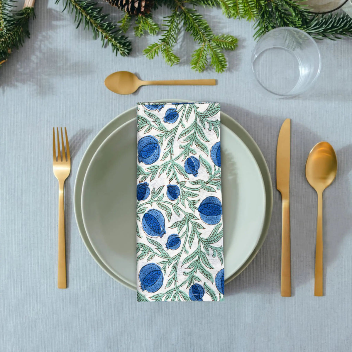 Table setting with a patterned napkin, green leaves, and gold cutlery on a gray tablecloth.