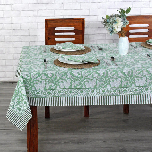 Dining table set with a green and white patterned tablecloth, wooden chairs, and a vase with flowers.