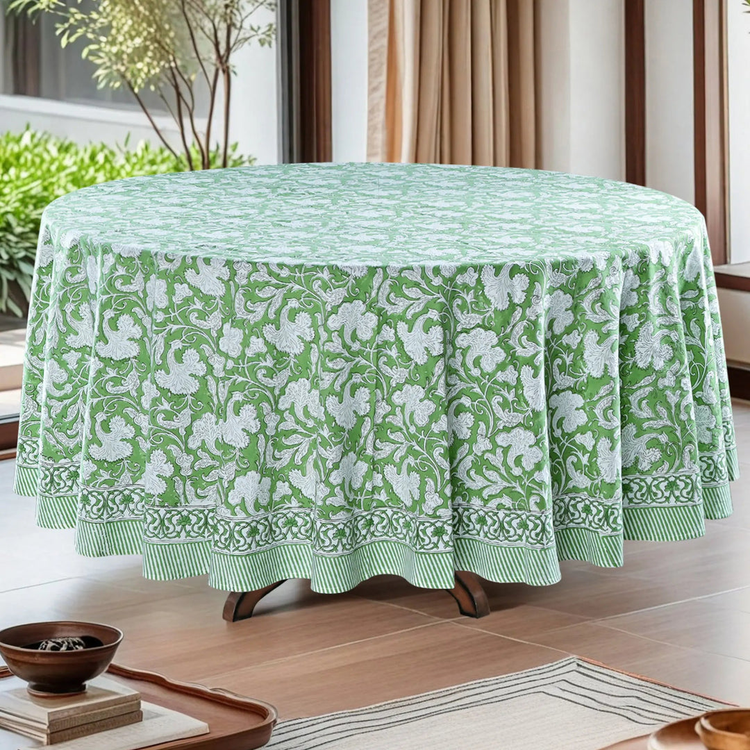 Green floral patterned tablecloth on a round table in a room with plants and curtains.