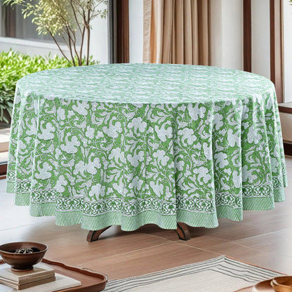 Green floral patterned tablecloth on a round table in a room with plants and curtains.