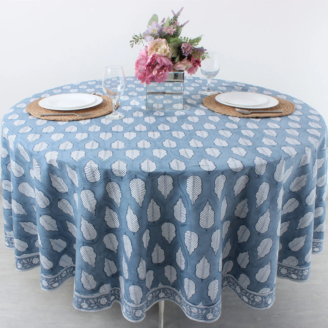 Round table with a blue patterned tablecloth, white plates, and floral centerpiece on a light background