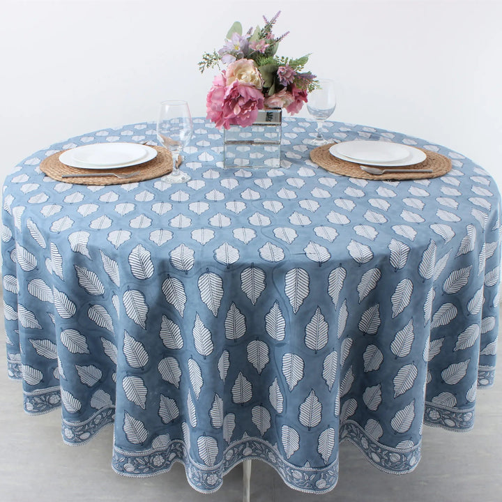 Round table with a blue patterned tablecloth, white plates, and floral centerpiece on a light background