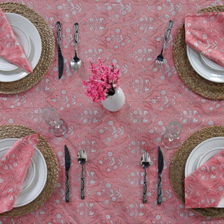 Dining table setting with pink floral tablecloth, plates, cutlery, and napkins.