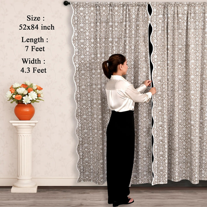Person adjusting a long curtain with floral pattern, next to a small table with flowers.