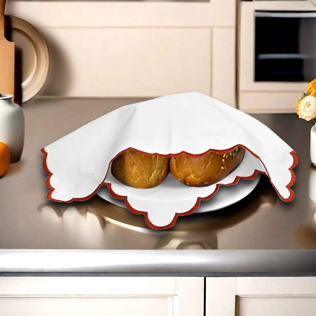Two baked goods on a white plate with a red rim, covered by a white cloth on a kitchen counter.